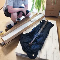 Mężczyzna rozpakowuje czarną katana na drewnianej skrzyni w pokoju.
